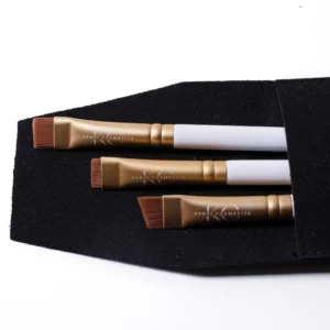 Set of three Kensi Cosmetics angled brushes with gold ferrules and white handles, neatly placed inside a black fabric pouch for professional brow styling